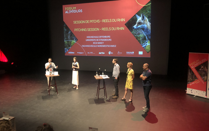 Moderierende und Jury auf der Bühne vor der Leinwand auf der die Pitches angekündigt werden