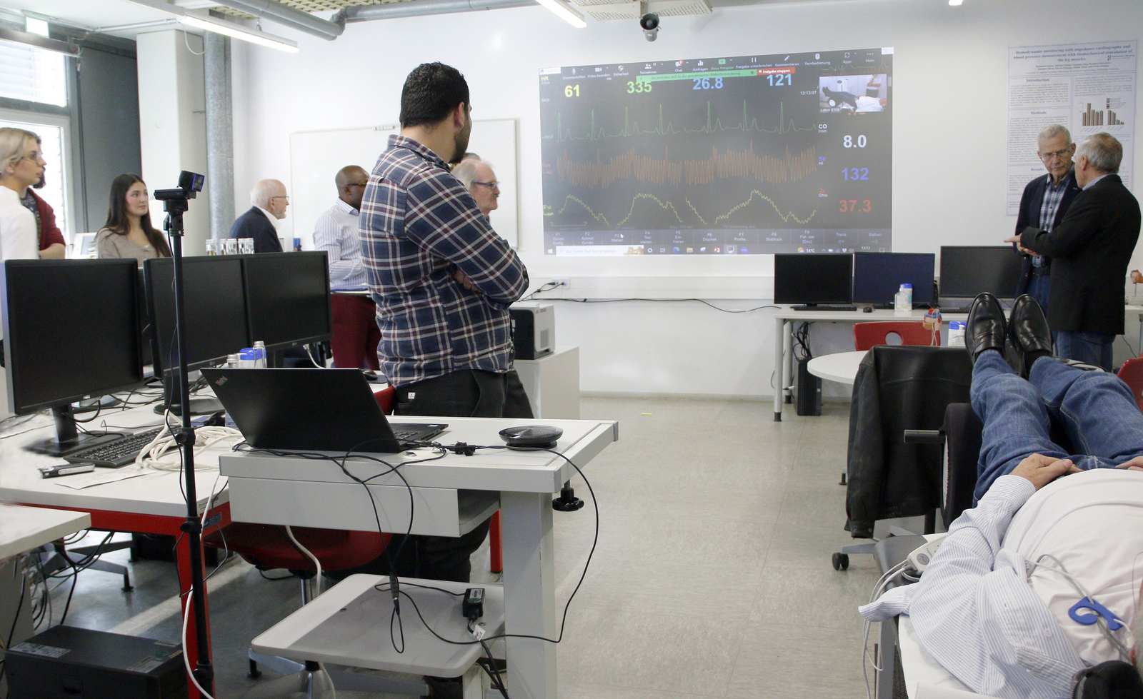 Eine Person liegt auf einer Liege während die Waden biomechanische stimuliert werden. Die anderen Anwesenden verfolgen auf einem Bildschirm die Veränderungen der gemessenen Herzparameter.
