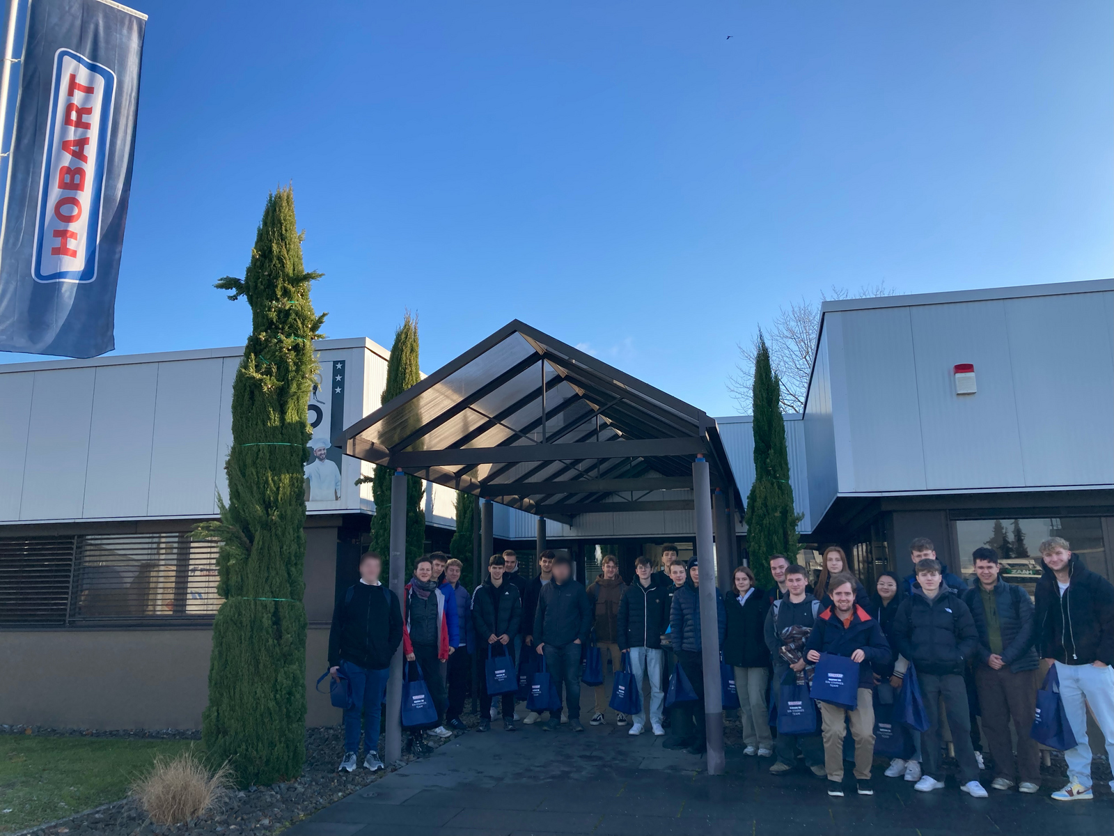 startING Studierende stehen als Gruppe vor dem Haupteingang von Hobart bei blauem Himmel. Links weht eine Hobartfahne im Wind.
