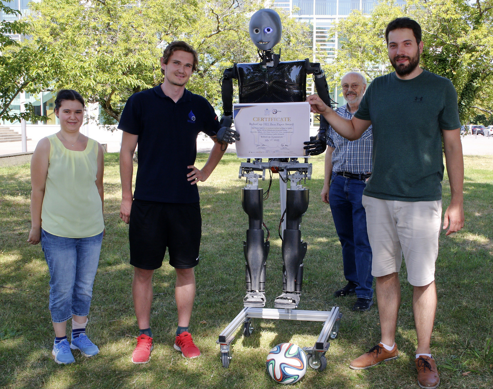 Sweaty steht umrahmt von Teammitgliedern auf einer Wiese, vor sich ein Fußball und hält in seinen Händen das Zertifikat 