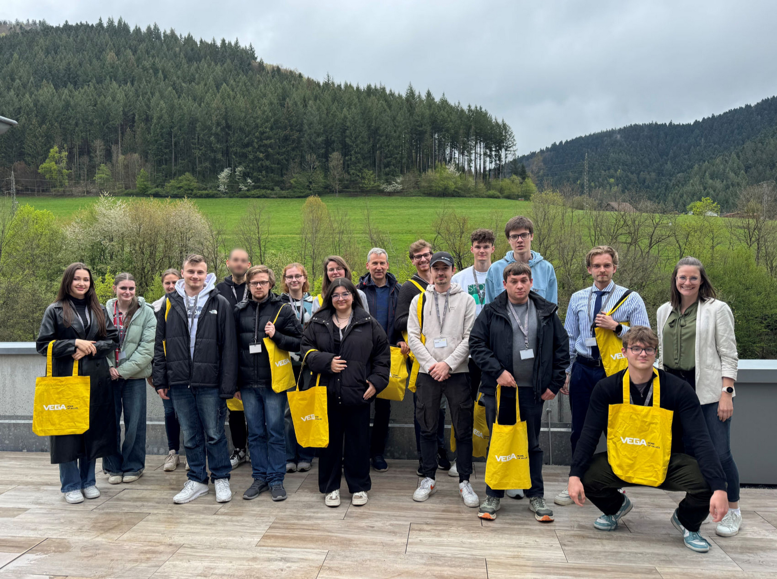 Exkursion zu VEGA im Sommersemester 2025 Gruppe Studierender steht mit gelben VEGA Taschen auf einer Terrasse, im Hintergrund sieht man Wald und Wiesen.