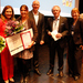EinE gruppe von chic gekleideten Menschen zwei Frauen haben Blumensträuße in den Händen, die eine zusätzlich noch eine Urkunde und einen Scheck 