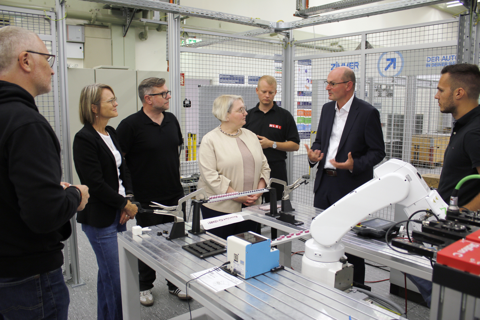 Eine Gruppe von Menschen steht um einen Tisch mit einerm Roboterarm herum, ein Mann erklärt etwas 