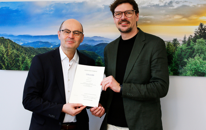 Zwei Männer in Anzügen stehen vor einem Schwarzwaldfoto und halten eine Urkunde zwischen sich