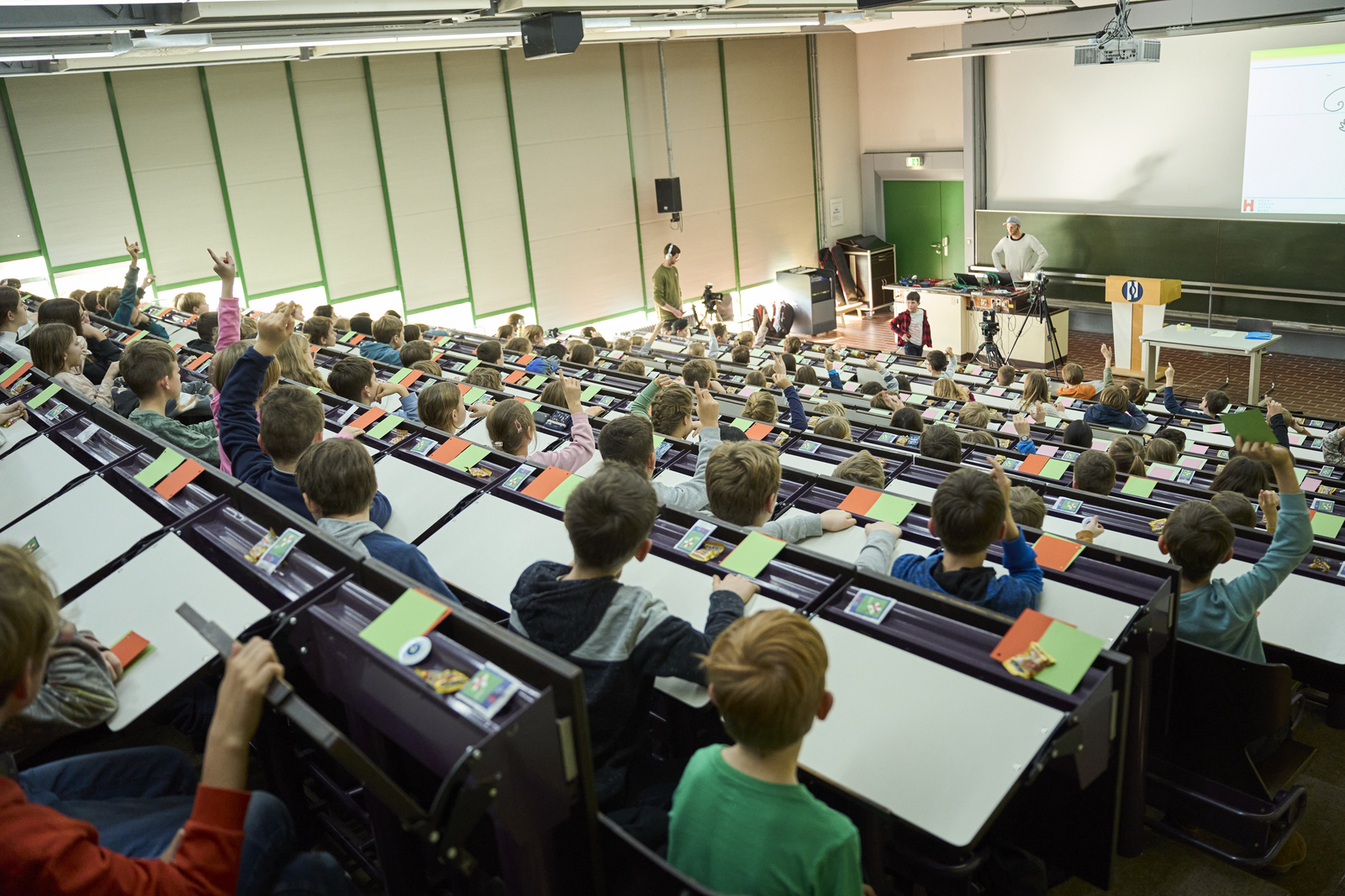 Kinder, die während einer Vorlesung im Saal sitzen. Viele davon melden sich.