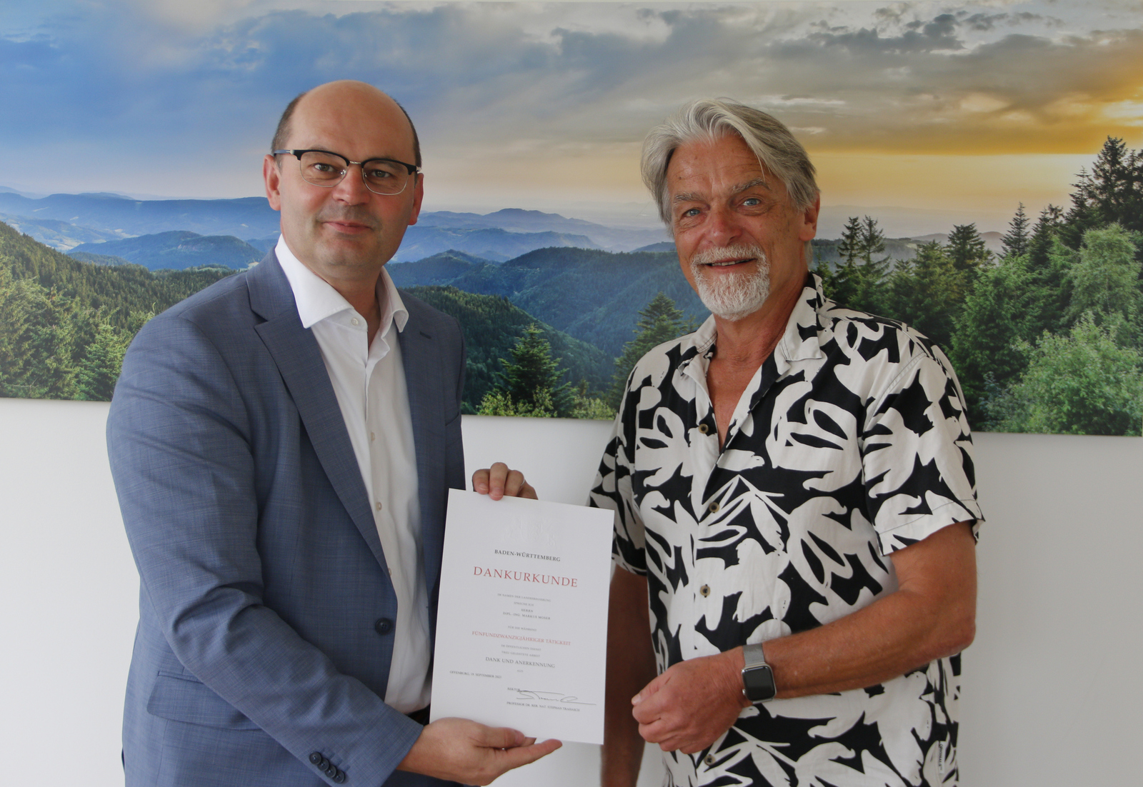 Stephan Trahasch (links) überreicht Markus Moser (rechts) vor einem Schwarzwaldpanorama stehend die Urkunde