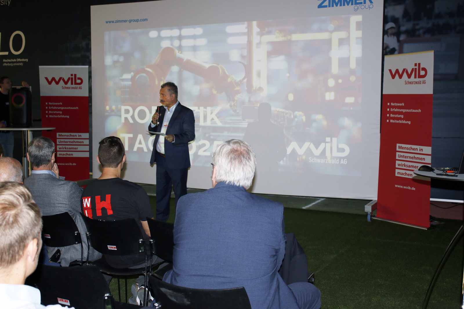 2025-07-15 wvib Robotik Tag Ein Mann steht vor einer Leinwand vor sitzenden Zuhörenden und spricht ins Mikrofon
