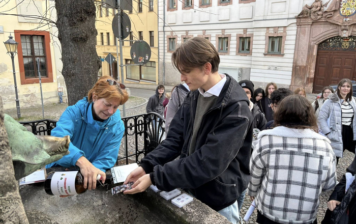 Eingruppe von menschen an einem Brunnen. Eine Frau gießt etwas aus einer Flasche über eine Spielkarte, die ein junger Mann hält