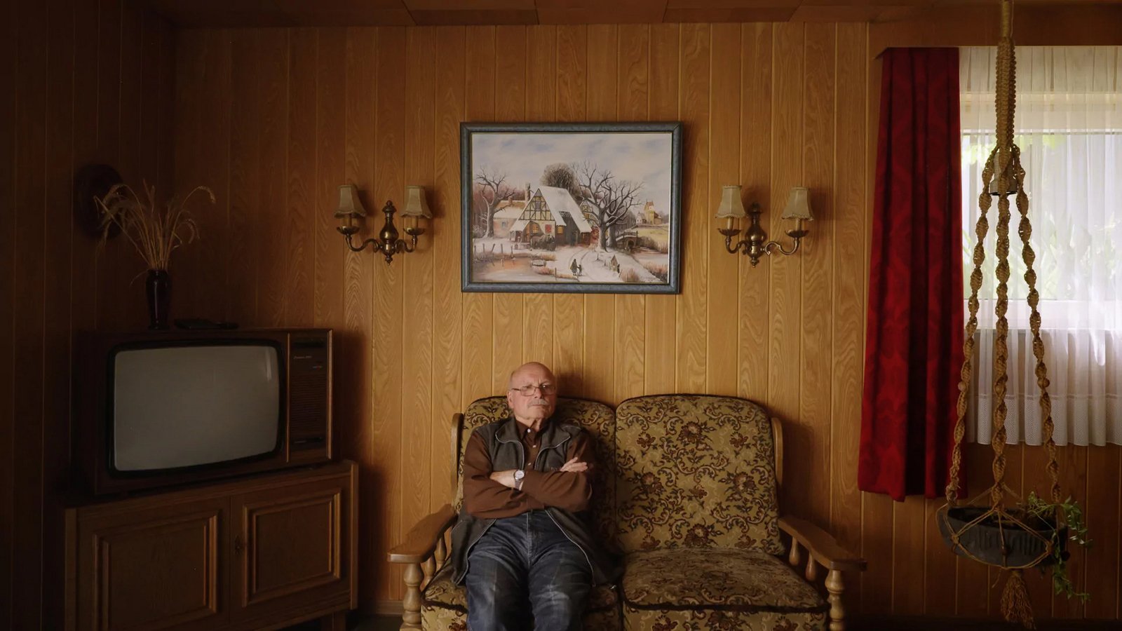 Eine alter Mann sitzt in einem altmodisch eingerichteten Wohnzimmer unter einem Bild auf einem Sofa