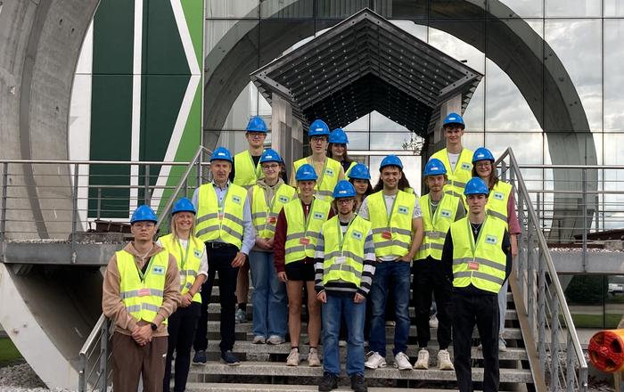 startING Studierende stehen in blauen Helmen und neongelben Warnwesten bekleidet vor dem Firmeneingang auf einer Treppe bei Herrenknecht