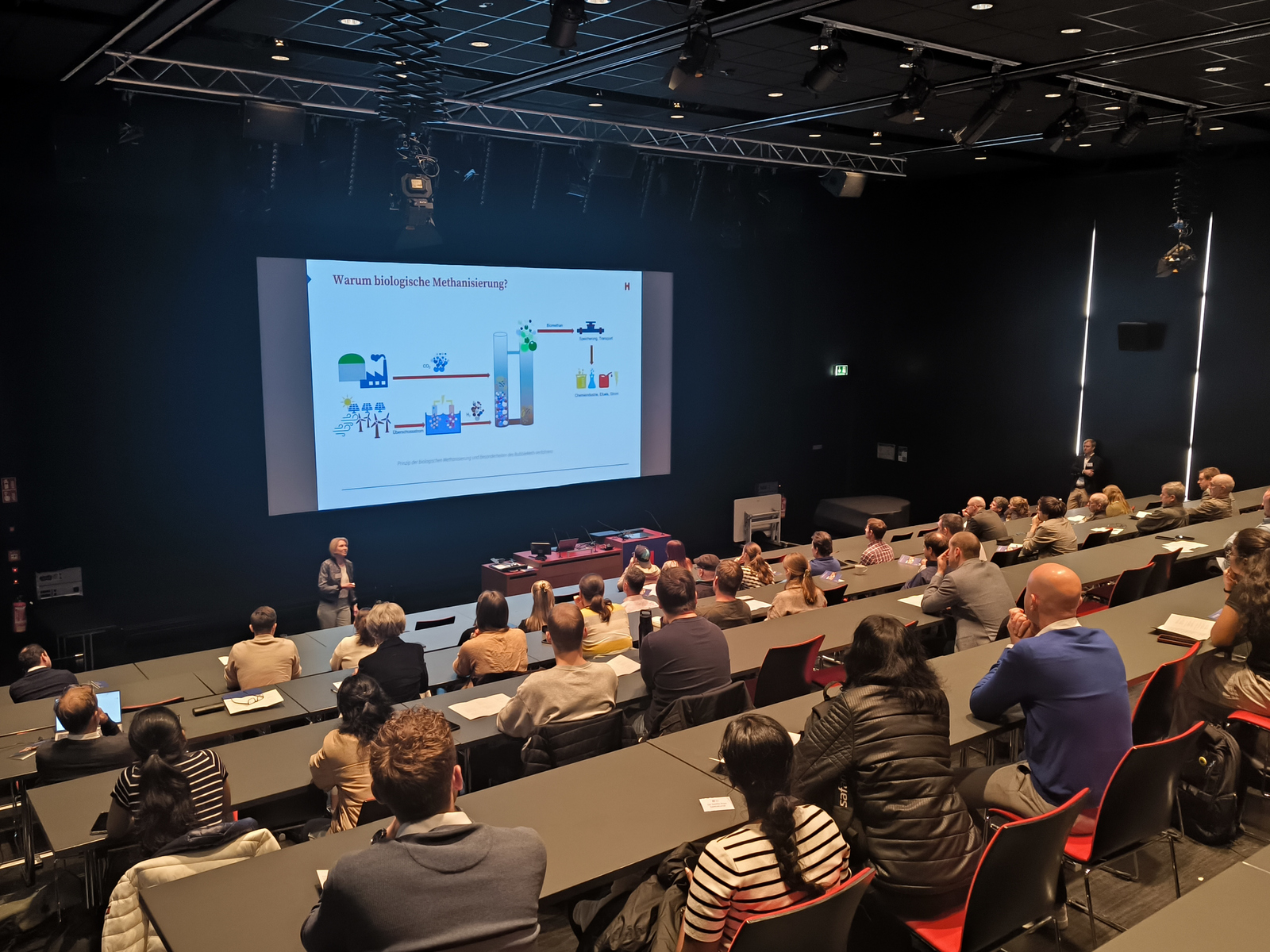 Eine Frau steht in einem Hörsaal vor einer Leinwand neben einem Rednerpult und spricht zu sitzenden Zuhörenden