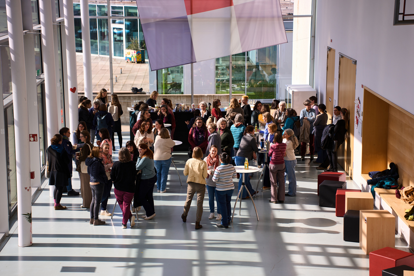 Frauen stehen in einem Foyer mit großen Fensterflächen durch die Sonne scheint zusammen und unterhalten sich.