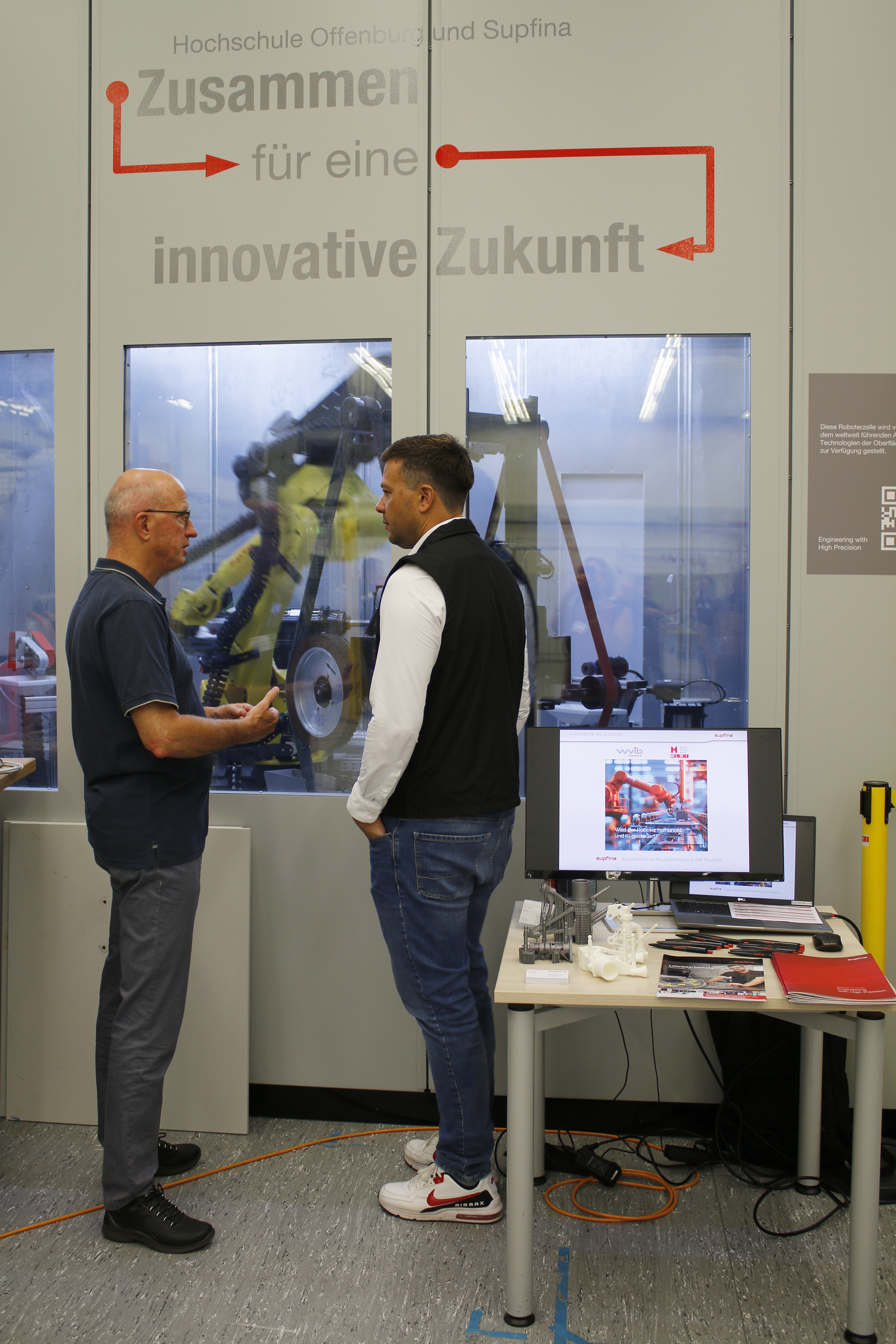 2025-07-15 wvib Robotik Tag Zwei Mäner stehen vor einem Fenster, dahinter arbeitet ein Industrieroboter