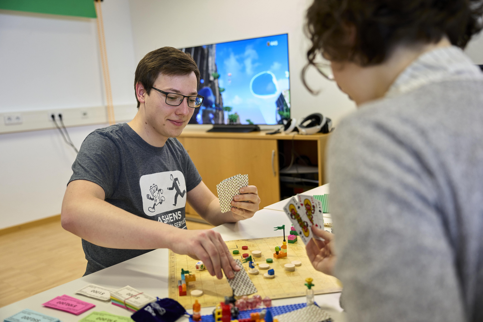 jungen Menschen sitzen an einem Tisch mit einem Brettspiel, Figuren und Karten im Hintergrund ist ein Bildschirm zusehen