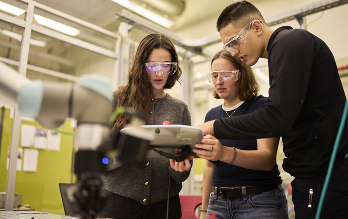 Zwei junge Frauen und ein junger Mann mit Schutzbrillen stehen vor einem Roboterarm und schauen auf ein Tablet zu dessen Bedienung