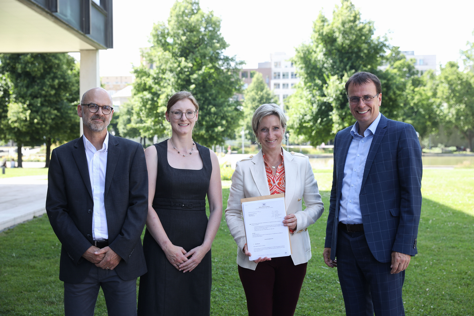 Prof. Dr. Tobias Hagen, Prof.in Dr. Simone Braun, Ministerin Dr. Nicole Hoffmeister-Kraut und Kultus-Staatssekretär Volker Schebesta (von links) stehen auf dem Rasen vor dem Ministerium. Die Ministerin hat den Bewilligungsbescheid in den Händen