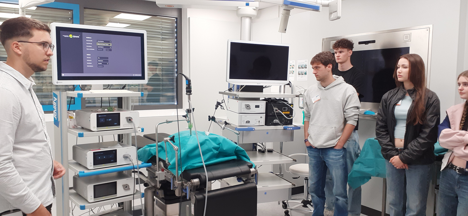 Vier startING Studierende stehen rechts im Bild in einem Operationsraum und schauen auf die Geräte und Displays. Ein Mitarbeiter steht links bei einem Monitor.