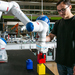Ein junger Mann mit schwarzem T-Shirt und schwarzer Hose steht in einem Labor an einem Roboterarm der bunte Bauklötze stapelt