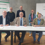 Drei Männer sitzen nebeneinander an einem Tisch und unterzeichnen die Kooperationsvereinbarung. Drei Menschen stehen hinter ihnen.
