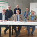 Drei Männer sitzen nebeneinander an einem Tisch und unterzeichnen die Kooperationsvereinbarung. Drei Menschen stehen hinter ihnen.