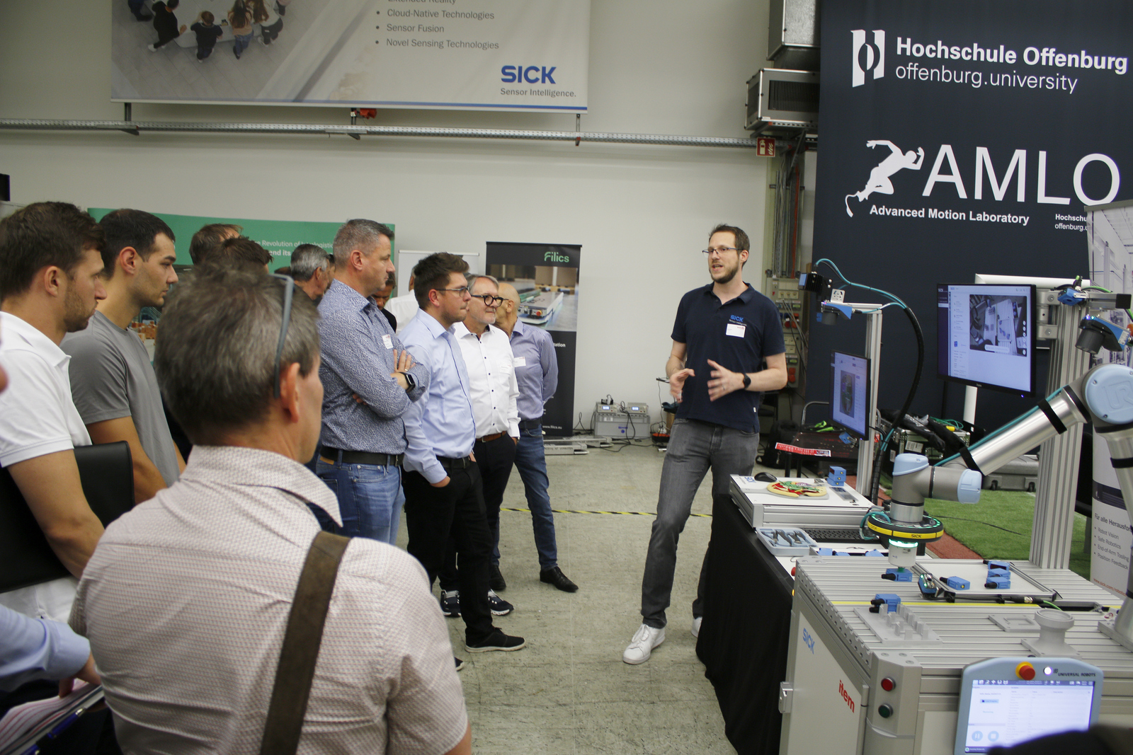2025-07-15 wvib Robotik Tag Ein Mann steht neben zwei kleinen Industrierobotern und erklärt den Umstehenden etwas