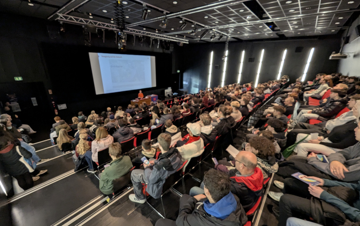 In einem Hörsaal sitzen zahlreiche jungen Menschen und hören eienr Frau zu, die vorn vor einer Leinwand an einem Rednerpult steht 