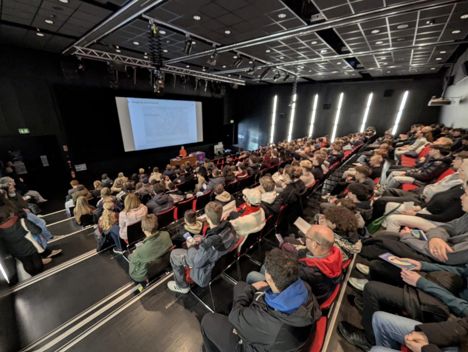In einem Hörsaal sitzen zahlreiche jungen Menschen und hören eienr Frau zu, die vorn vor einer Leinwand an einem Rednerpult steht 