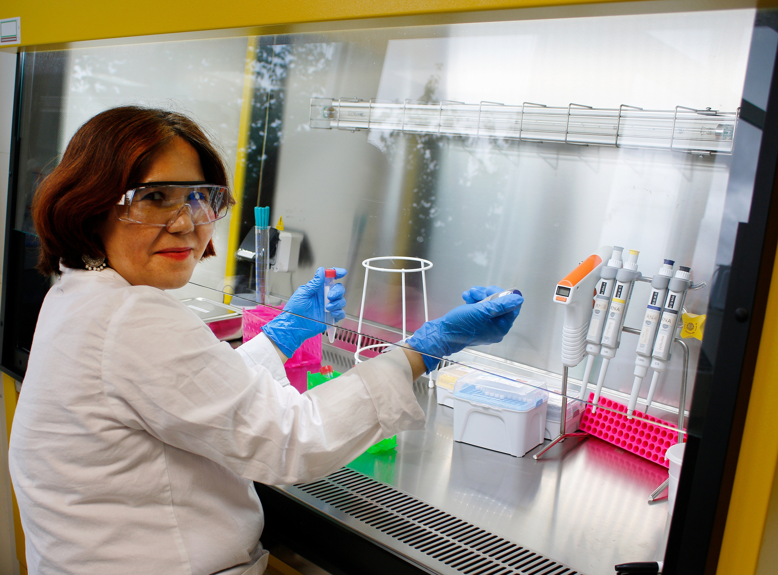 Eine Frau im weißen Kittel mit Laborbrille und Handschuhe sitzt in einem Labor und arbeitet mit Pipette und Reagenzglas