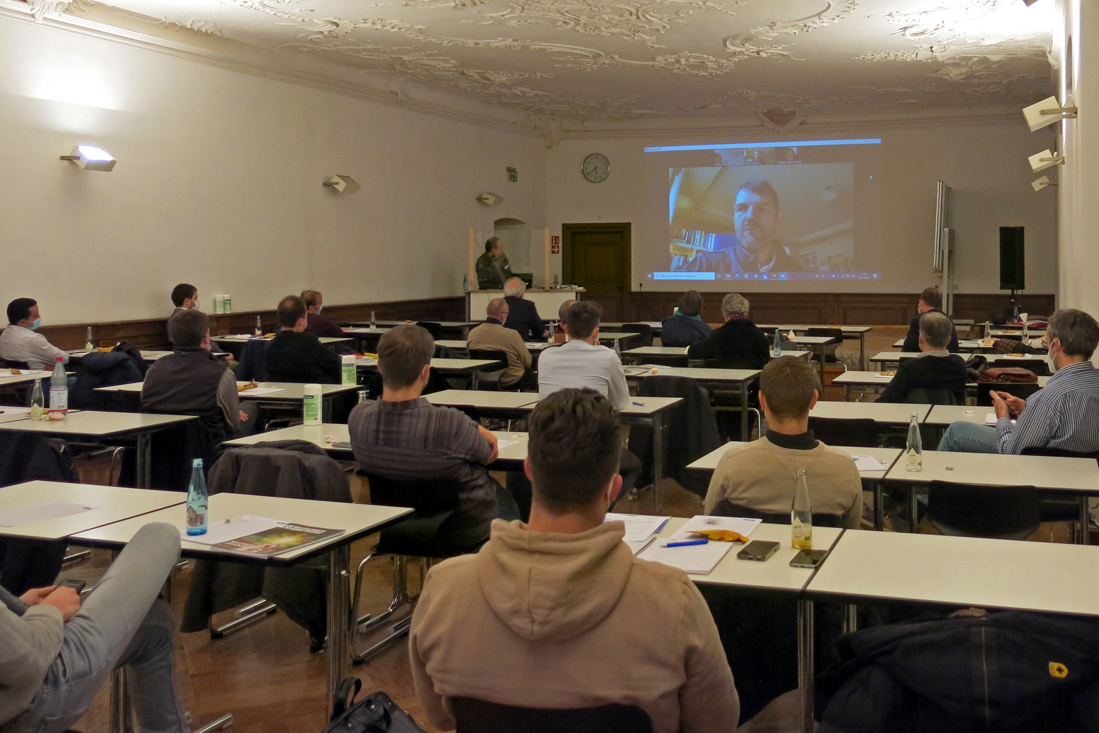 Im Barocksaal des Klosters Gengenbach sitzenden die Zuhörenden vor einer leinwand auf der Live-Übertragung des Vortrags von Dr. Mario Herger aus dem Silicon Valley zu sehen ist. 