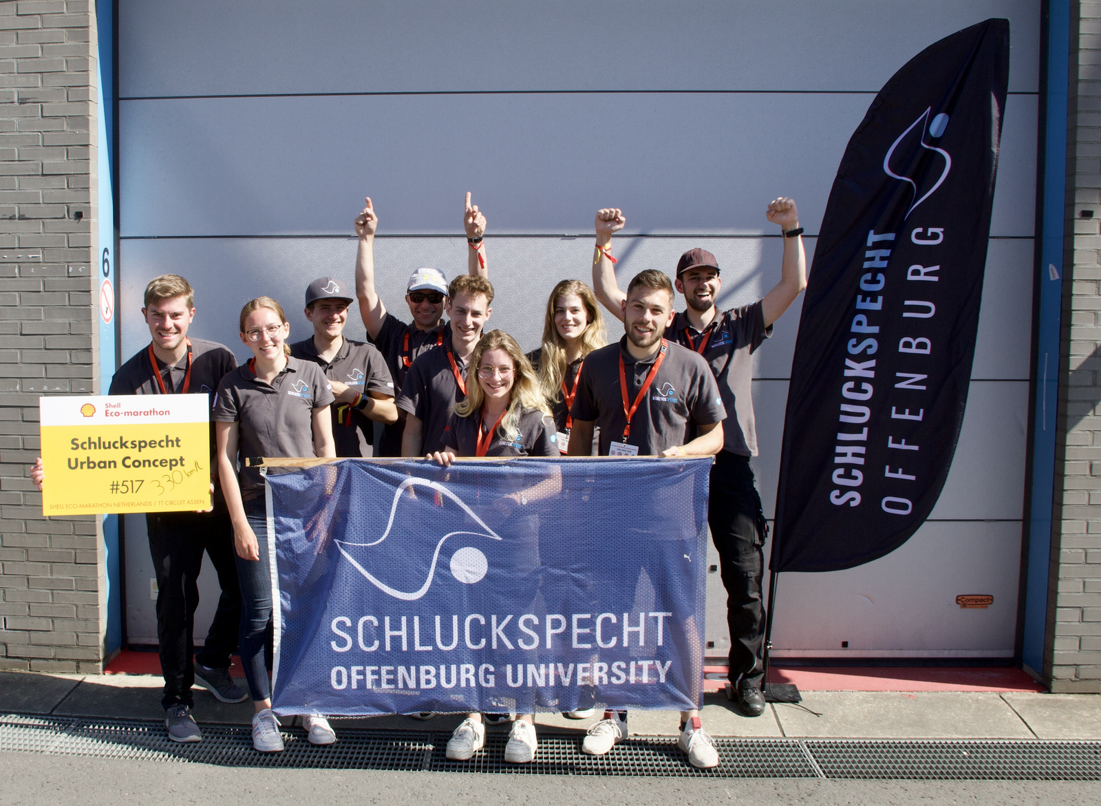 Das jubelnde Team mit Teamfahnen und einem Startschild auf dem handschriftlich die Kilometerleistung mit einem Liter Diesel notiert ist vor einem Garagentor 