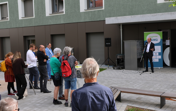 Ein Mann steht vor einem Gebäude und spricht zu Menschen an Stehtischen