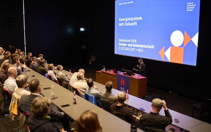 Ein Mann steht in einem Hörsaal vor einer Leinwand mit einer Präsentation an eine Rednerpult und spricht zu sitzenden Zuhörenden 