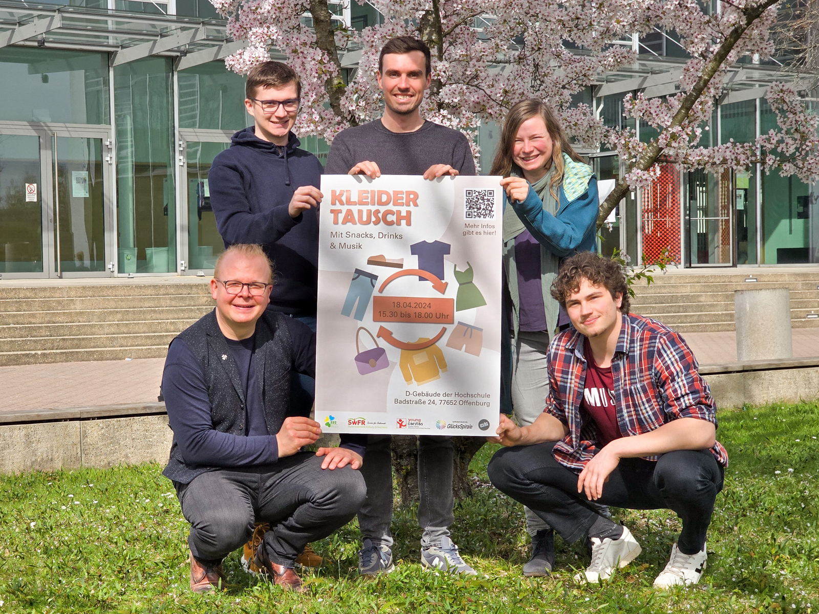 Die Veranstalter knien und stehen auf der Wiese vor Gebäude D und halten Plakat der Kleidertauschbörse hoch.