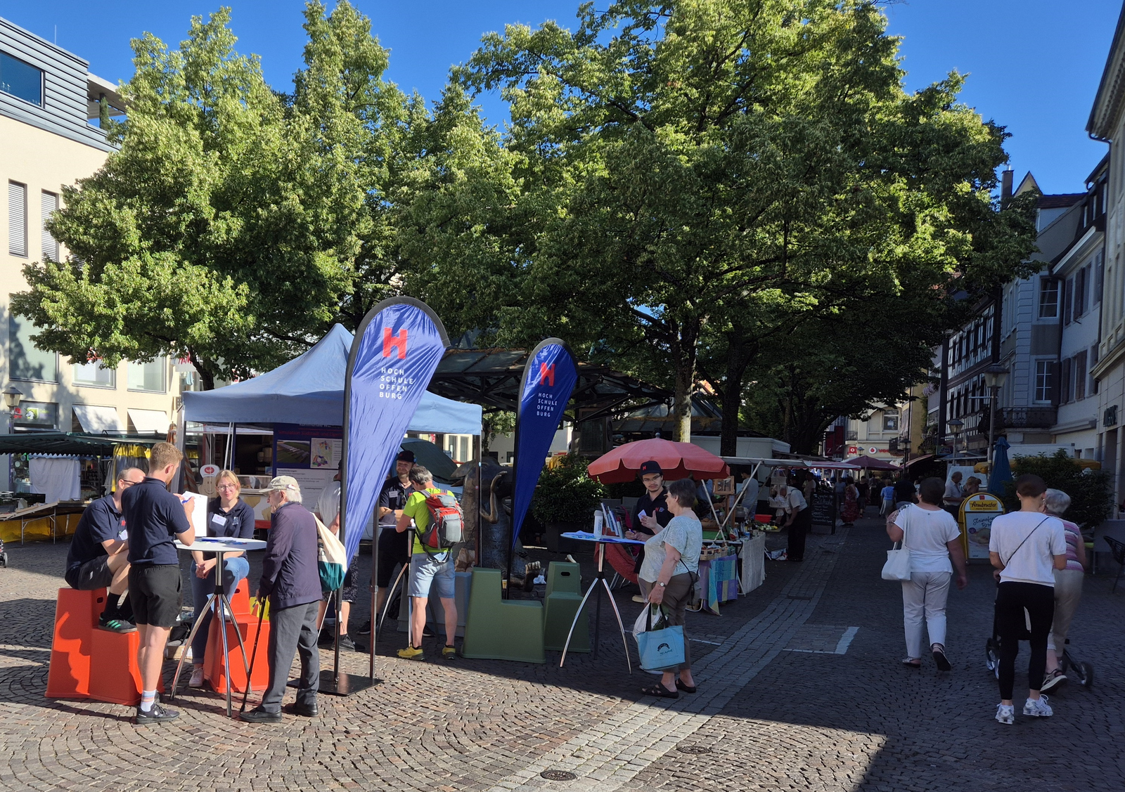 Marktbesucherinnen und -besucher werden an einem blauen Hochschulpavillonzelt und Stehtischen von Studierenden befragt