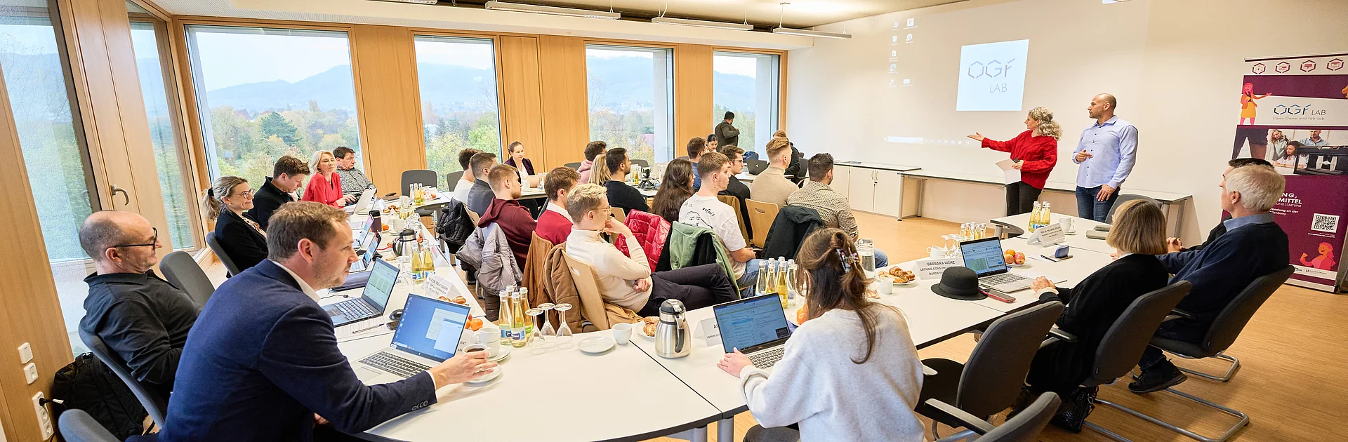 In einem Konferenzraum sitzen viele Leute im Anzuig und auch viele Studierende. Zwei Personen präsentieren neben einer Beamerfläche.
