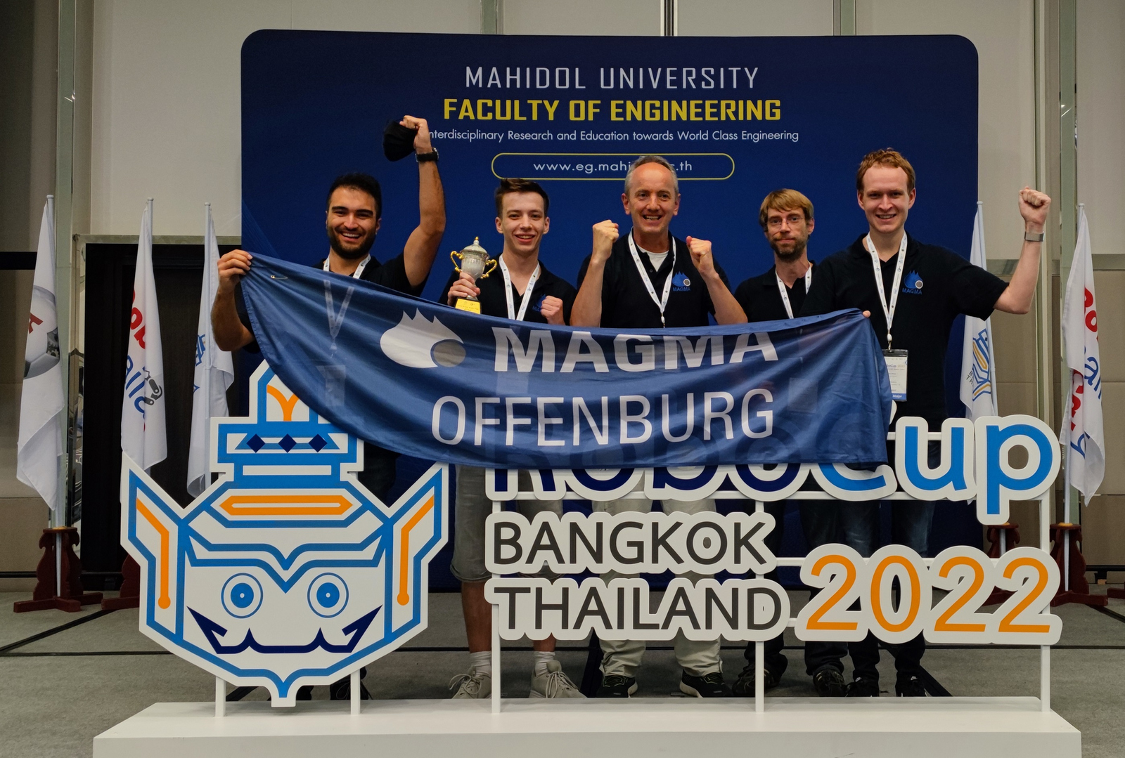 Die Teammitglieder mit dem Pokal und einer Team Fahne hinter dem RoboCup Logo 