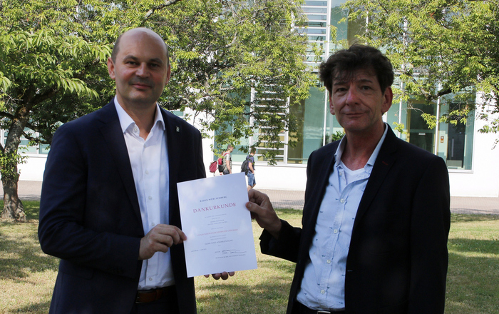 Rektor Prof. Dr. Stephan Trahasch (links) und Prof. Dr. Gerhard Kachel (rechts) stehen bei der Urkundenübergabe auf dem Campus unter Bäumen.