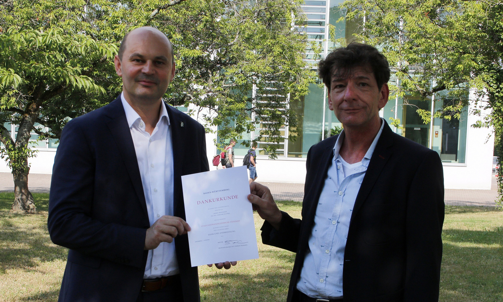 Rektor Prof. Dr. Stephan Trahasch (links) und Prof. Dr. Gerhard Kachel (rechts) stehen bei der Urkundenübergabe auf dem Campus unter Bäumen.