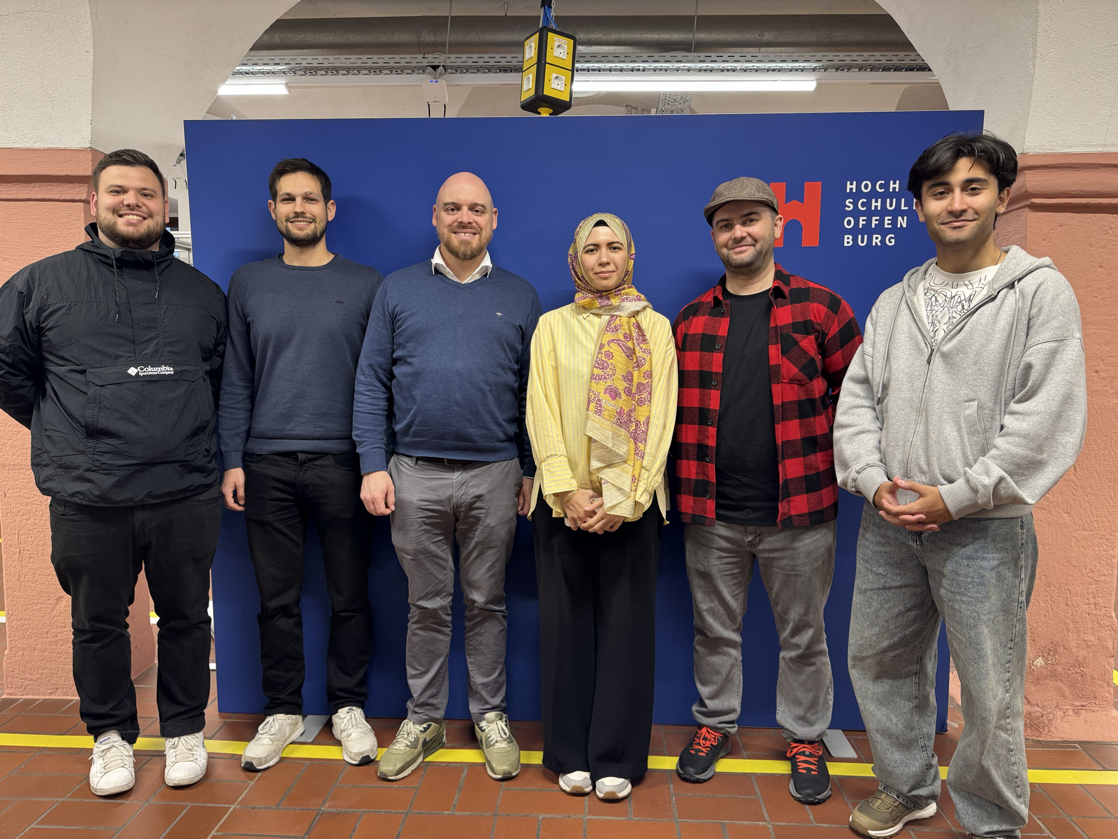 Sechs Personen stehen nebeneinander vor einer blauen Wand mit dem Schriftzug 'Hochschule Offenburg'.