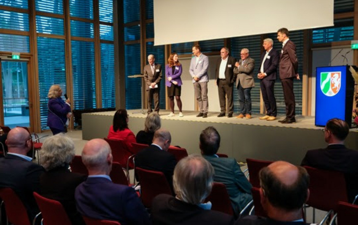 Eine Gruppe von Leuten steht auf einer Bühne vor sitzenden Zuhörenden