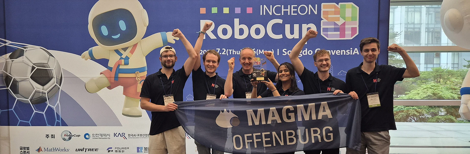 Gruppenfoto von Team Magma, wie sie einen Banner mit ihrem Namen und den gewonnenen Pokal hochhalten