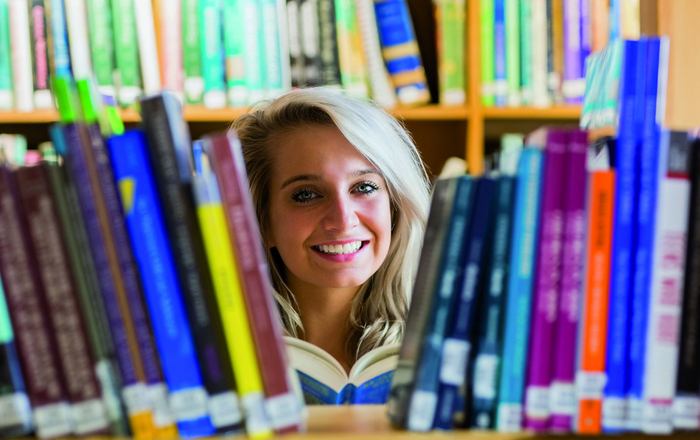 Frau hält ein blaues Buch in der HAnd und durch schat durchen eine Lücke zwischen Büchern in einem Regal hindurch