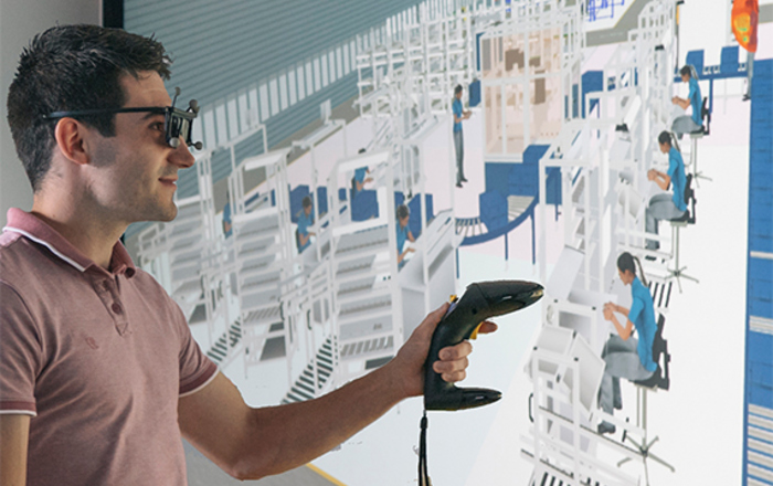 Ein junger Mann mit einer VR-Brille vor einem Bildschirm auf dem das gezeichnete Innere einer Fabrik zu sehen ist