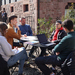 Eine Gruppe junger Leute sitzt draußen an einem Tisch mit Laptops und Unterlagen. Im Hintergrund stehen weitere junge Leute.