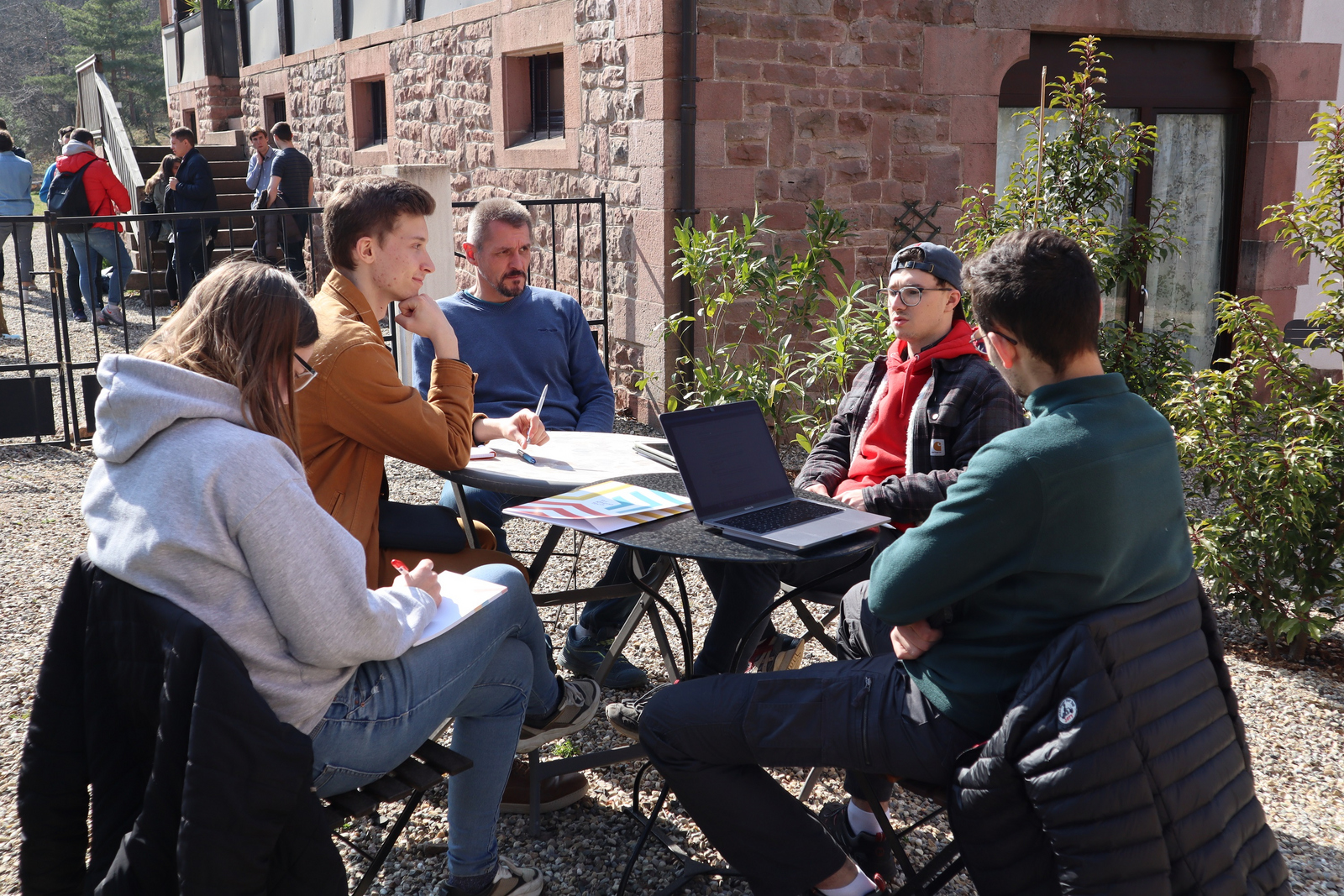 Eine Gruppe junger Leute sitzt draußen an einem Tisch mit Laptops und Unterlagen. Im Hintergrund stehen weitere junge Leute.