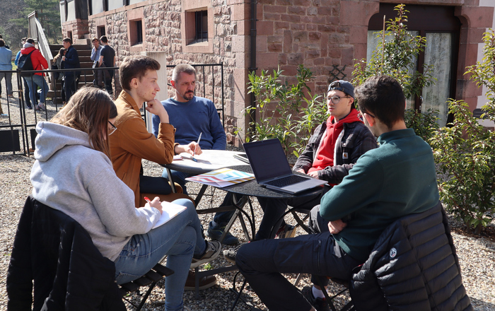Eine Gruppe junger Leute sitzt draußen an einem Tisch mit Laptops und Unterlagen. Im Hintergrund stehen weitere junge Leute.