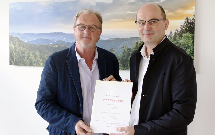 Heiner Behring (links) und Stephan Trahasch (rechts) stehen mit der Urkunde vor einem Schwarzwaldbild 