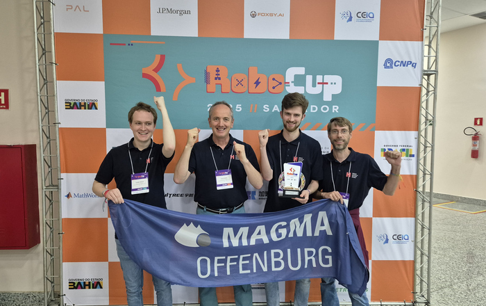 Vier Männer stehen mit Teamfahne und Pokal vor einer RoboCup Wand und jubeln