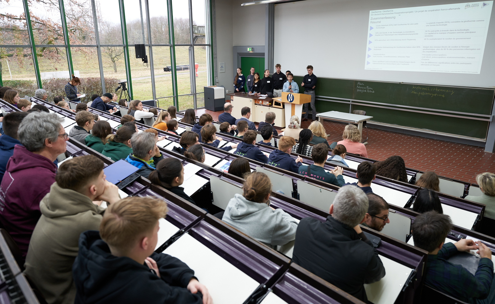 Schülergruppe steht vor sitzenden Jugendlichen im Hörsaal und präsentiert Geothemieprojekt