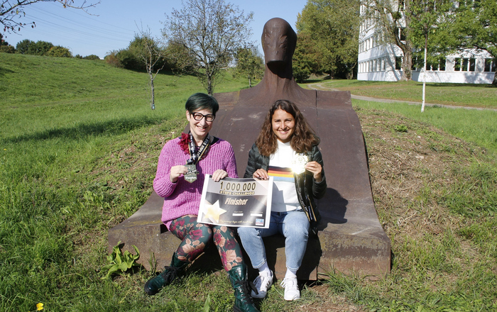 Cornelia Herde und Stella Schneider (von links) sitzen auf der liegenden Eisenskulptur an der Hochschule und halten ihre Finisher-Medaillen und eine Urkunde in die Kamera